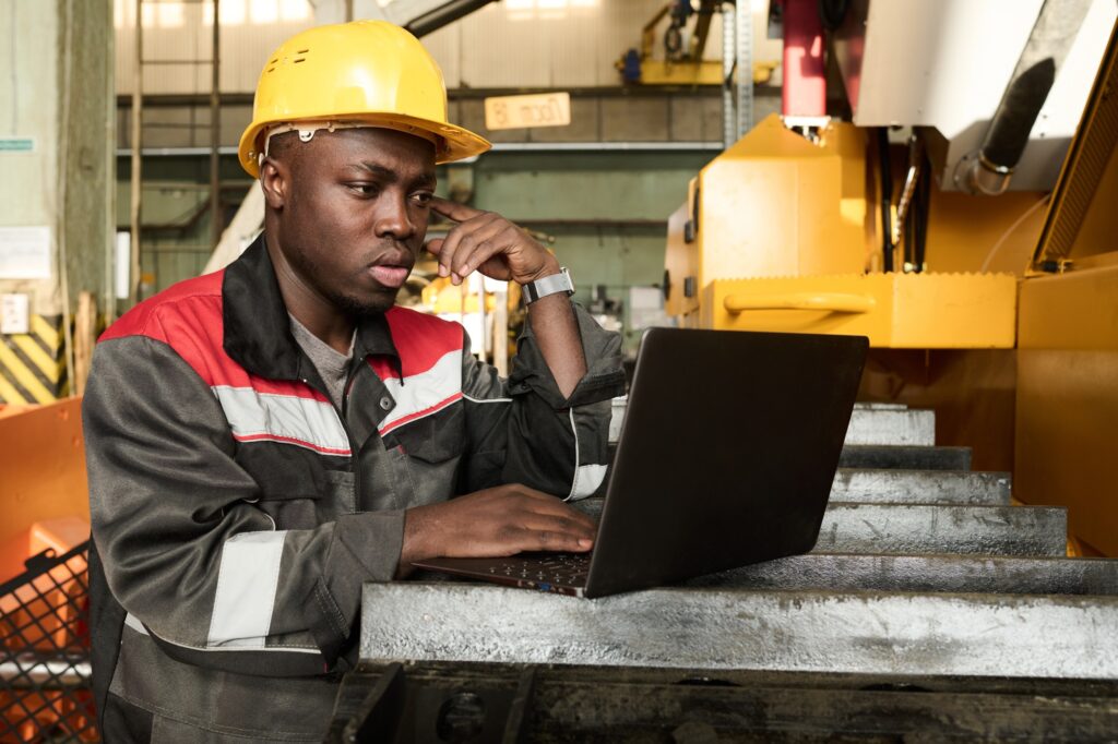 Young pensive worker of industrial plant using laptop by workplace